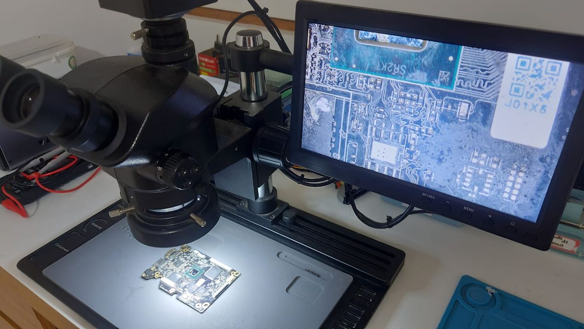 Microscópio trinocular sobre a bancada da Abratek, com uma placa-mãe de notebook ampliada no monitor ao lado